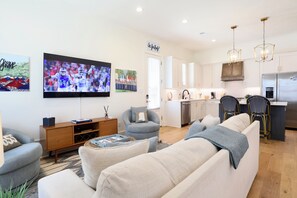 En moderne stue med et fladskærms-tv, en sofa, et sofabord og et spiseområde med et køkken i baggrunden.