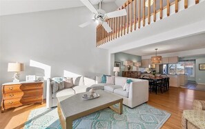 A spacious living room with a high ceiling, a wooden staircase, and a kitchen area visible in the background. - Pebble Paradise - Easy Walk to the Beach & Town (Bethany Beach)