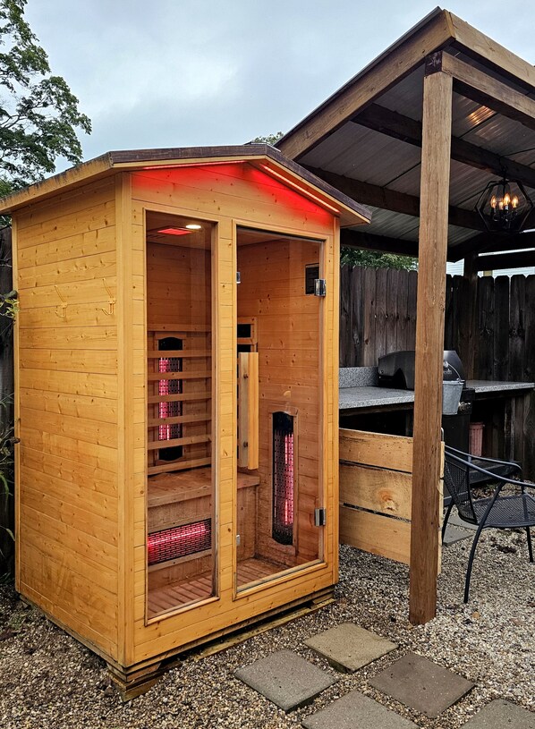 A wooden sauna structure with a glass door, a bench inside, and a grill outside.