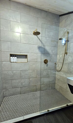 A shower area with a glass door, a handheld showerhead, and a wall-mounted showerhead. There is a shelf with toiletries and a hexagonal patterned floor.