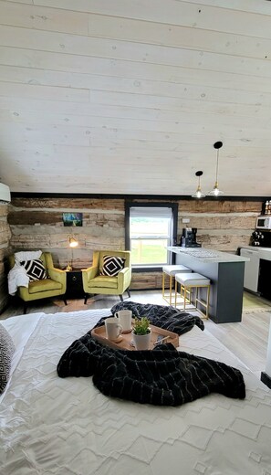 A cozy room with a bed, two chairs, a coffee table, and a kitchen area with a window.