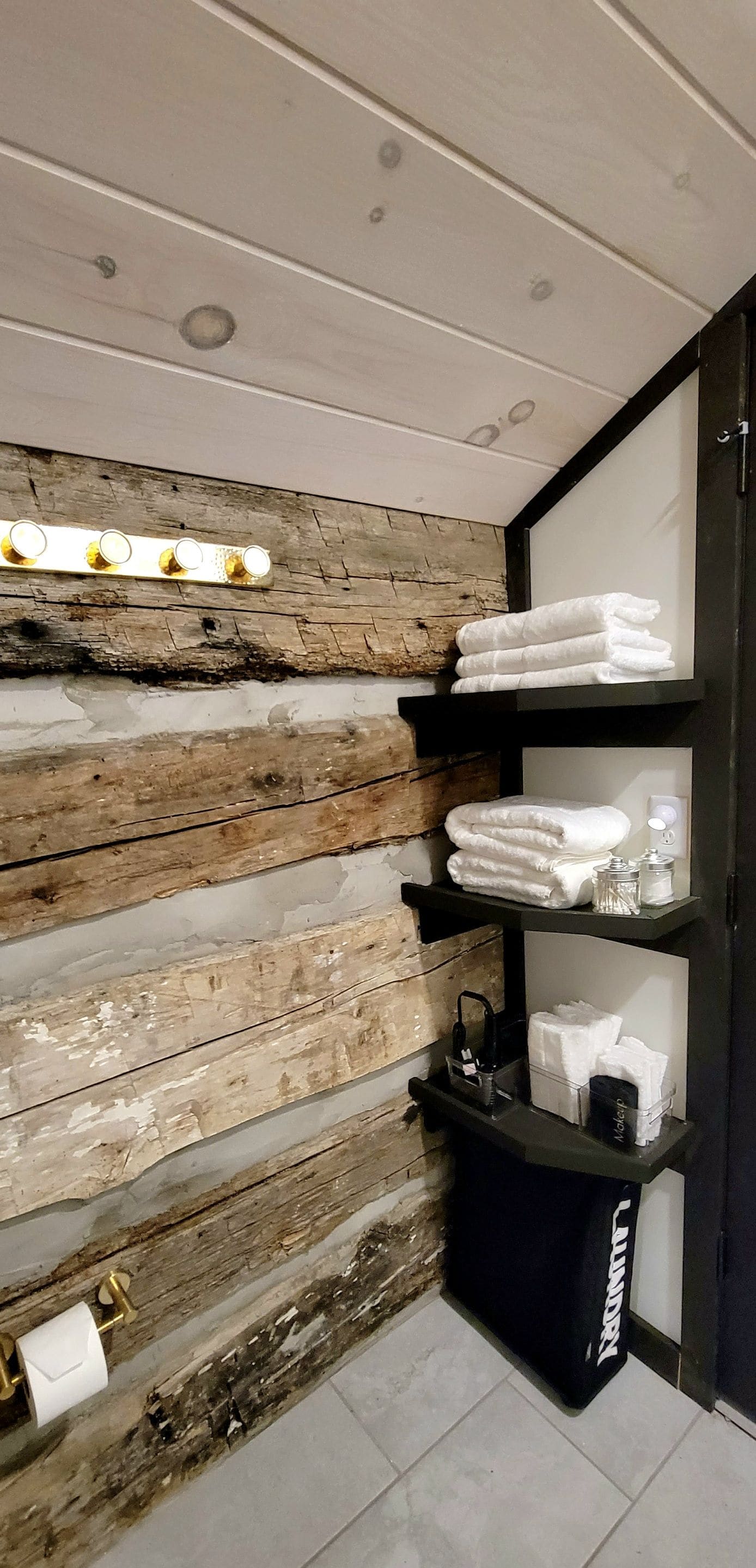 A bathroom with a rustic wooden wall, a black shelf with folded towels, and a black bag on the floor.