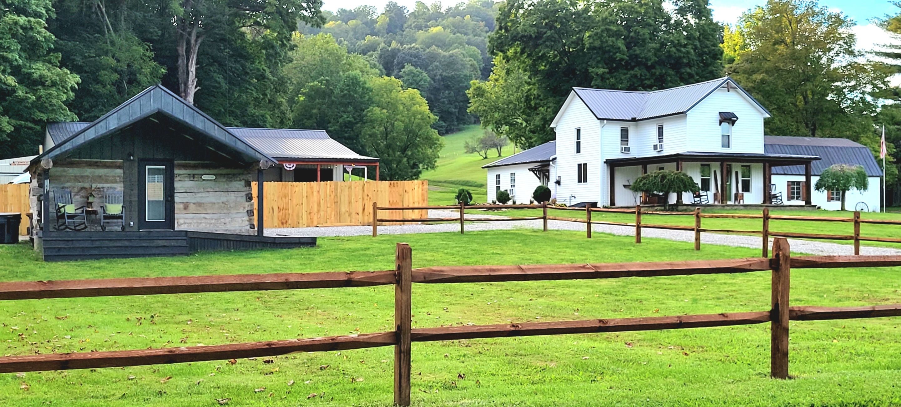 A rustic cabin with a porch and a white building with a porch in a grassy area with trees in the background.