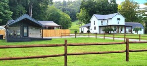 A rustic cabin with a porch and a white building with a porch in a grassy area with trees in the background.