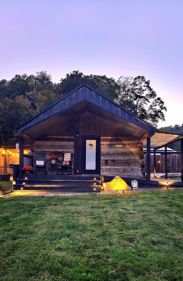 A rustic log cabin with a porch, a welcome sign, and a fire pit.