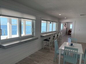 A room with a view of the sea, a dining area with chairs and a table, and a hallway leading to another room.