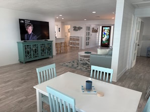 Una sala de estar moderna con televisor, mueble azul verdoso, sofá blanco y un comedor con mesa y sillas.