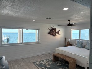 A bedroom with a bed, a bench, a ceiling fan, and a view of the water through the windows.