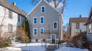 A two-story house with a grey exterior, a front door, and a small front yard with a snow-covered ground. - Walk to Brady St, 2-story upper flat, Sleeps 7! (Milwaukee)