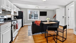A modern kitchen with white cabinets, a black refrigerator, and a stove. There is a black sofa with a red throw blanket and a small table with a vase. A TV is mounted on the wall above the sofa. - Walk to Brady St, 2-story upper flat, Sleeps 7! (Milwaukee)