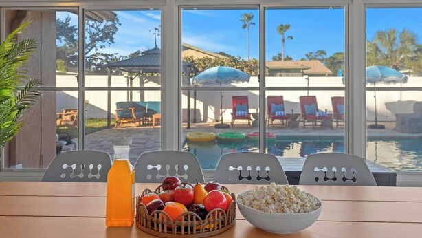 Mesa com cesta de frutas, garrafa de suco de laranja e tigela de pipoca, com vista para área da piscina com espreguiçadeiras e guarda-sóis.
