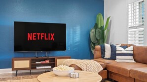 Uma sala de estar com uma televisão de tela plana grande exibindo "NETFLIX", um sofá de couro marrom, uma mesa de centro de madeira com um recipiente de pipoca e uma planta em vaso.