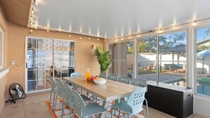 Uma sala de jantar com uma mesa comprida, cadeiras e uma cesta de frutas. Há um ventilador e uma planta na mesa.