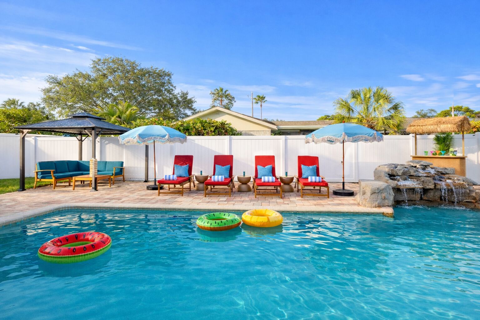A swimming pool with lounge chairs and umbrellas, a small waterfall feature, and a thatched-roof structure.