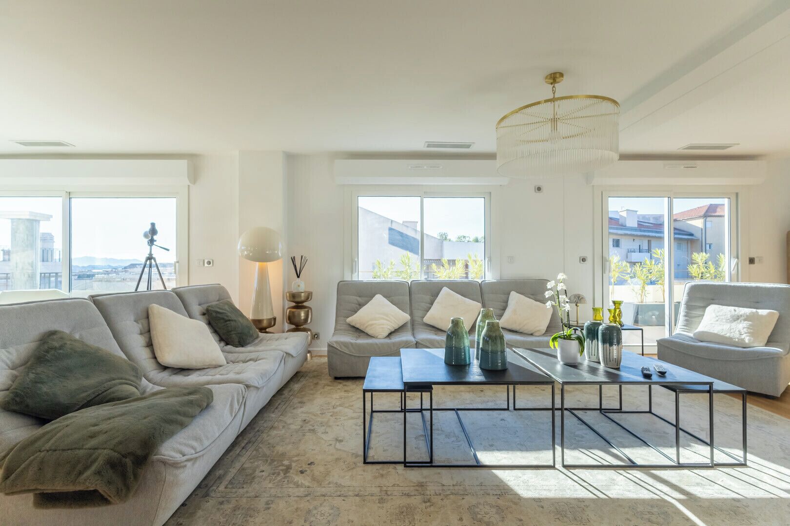 Ein geräumiges Wohnzimmer mit einer großen Couch, einem Couchtisch und einem Kronleuchter. Große Fenster bieten Blick auf die Stadt.