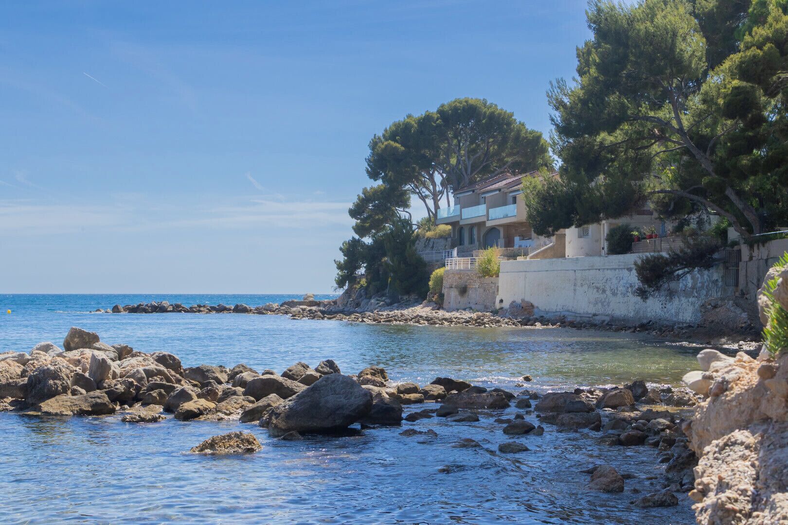 A coastal scene with a rocky shoreline, a building with balconies overlooking the sea, and trees.
