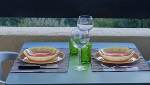 Une table avec deux assiettes, un verre et un verre vert, placée sur un balcon donnant sur une étendue d’eau et des arbres.