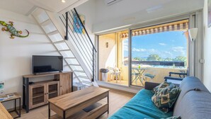 Un salon moderne avec un téléviseur, des meubles en bois, un canapé agrémenté de coussins à motifs, un escalier et un balcon avec des sièges d’extérieur.