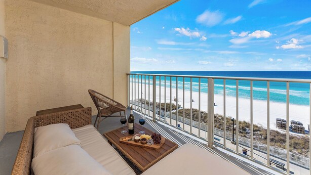 Een balkon met uitzicht op het strand, een rieten bank, een houten tafel met eten en drinken, en een stoel.