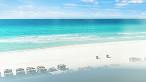 Een strand met helder turquoise water en een witte zandkust. Er zijn meerdere strandparasols en ligbedden zichtbaar.
