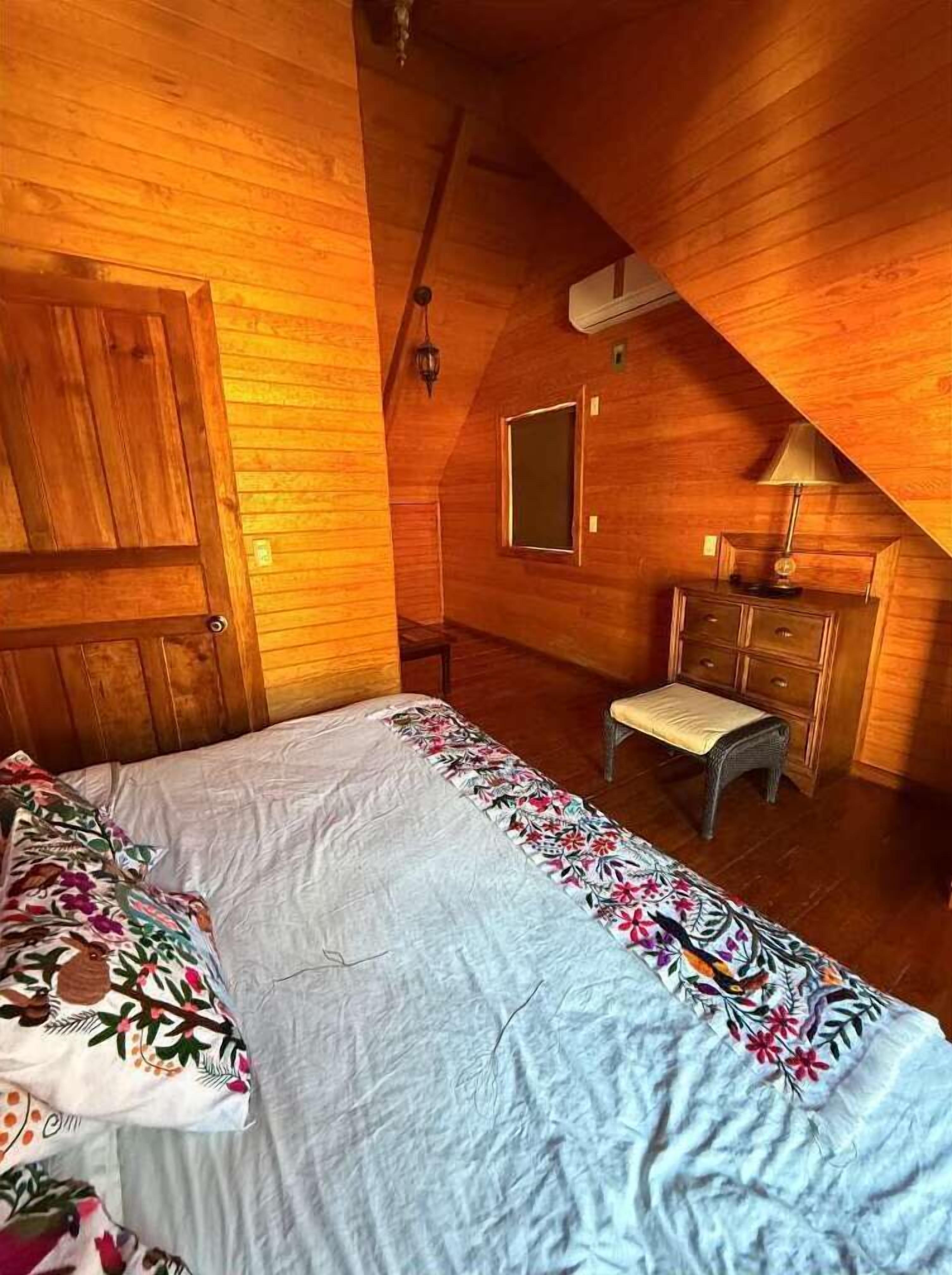 Un dormitorio con techo inclinado de madera, una cama con ropa de cama floral, un banco, un aparador y un espejo.