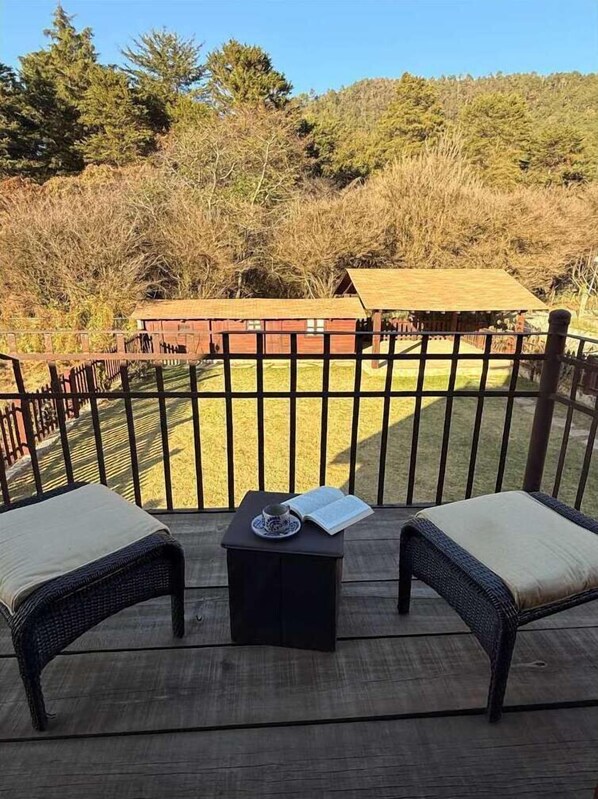A deck with two wicker chairs, a small table with a book and a cup, and a view of a building and trees in the background.