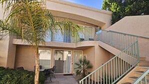 Un edificio de dos pisos con un balcón, una escalera y un patio con mobiliario exterior.