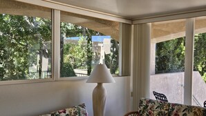 Una habitación con sofá y lámpara, ventanas amplias con vistas a zonas verdes y edificios.