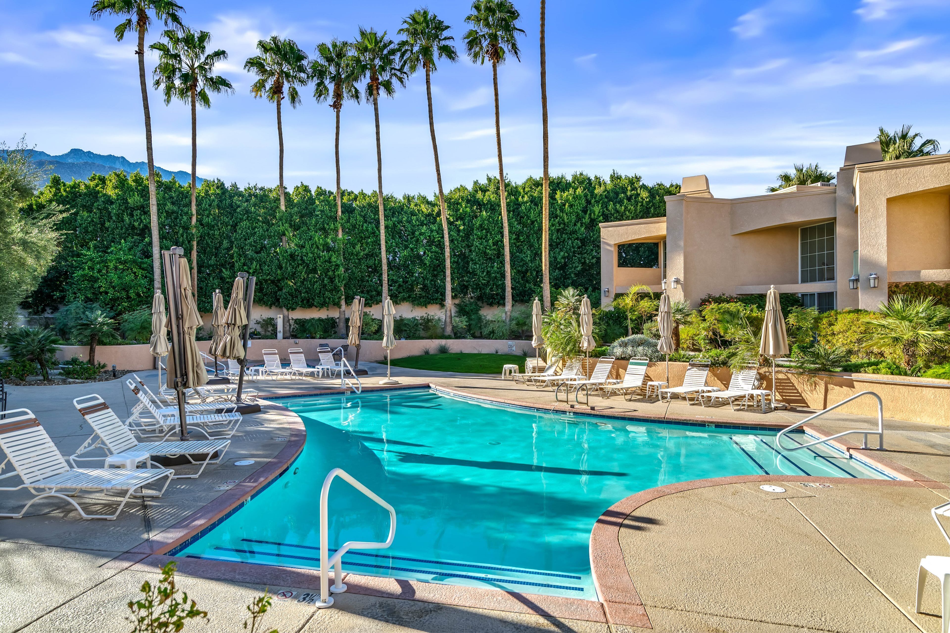 A swimming pool with lounge chairs and umbrellas, surrounded by palm trees and a building with a modern design.