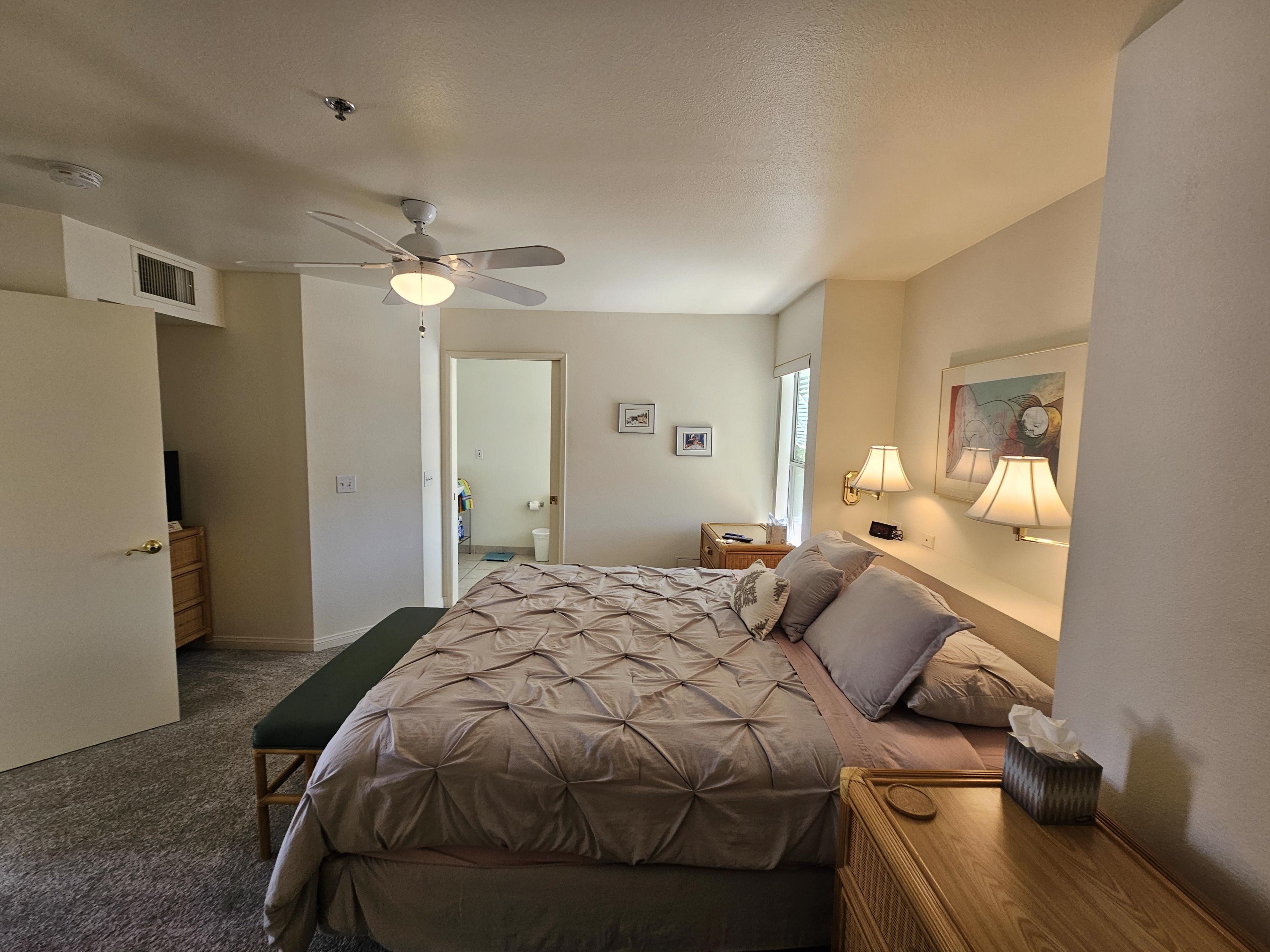 A hotel room with a bed, a bedside table, a lamp, a ceiling fan, and a door leading to another room.