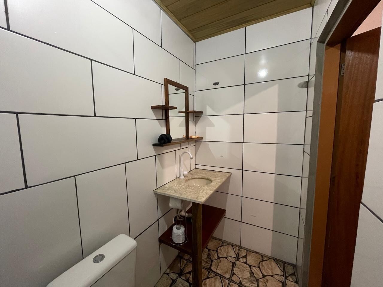 A small bathroom with white tiled walls, a beige sink, a wooden shelf, and a toilet.