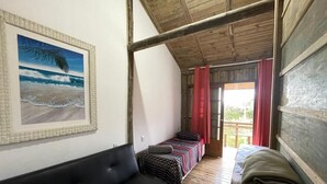 A room with a black sofa, a bed with a striped blanket, and a painting of a beach scene. - Cabin surrounded by nature, close to the beach and dunes. (Imbituba)