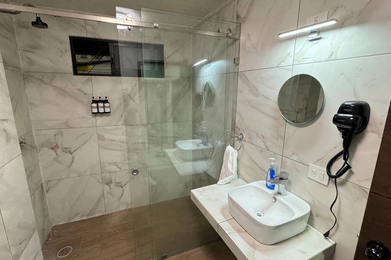 Ein modernes Badezimmer mit einer ein Glasfach raumteilenden Dusche, einem weißen Waschbecken mit Seifenspender und einem an der Wand befestigten Föhn.