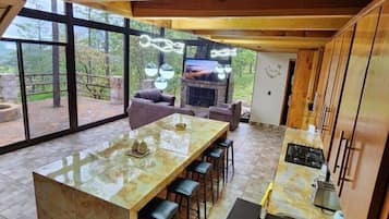 A modern kitchen with a large island, bar stools, a fireplace, and large windows offering a view of the outdoors.
