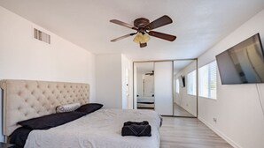 A bedroom with a tufted headboard, a ceiling fan, a bed with a blanket, a bedside table, and a large window. - The Pool House New 2-Story Home Near Strip (Las Vegas)