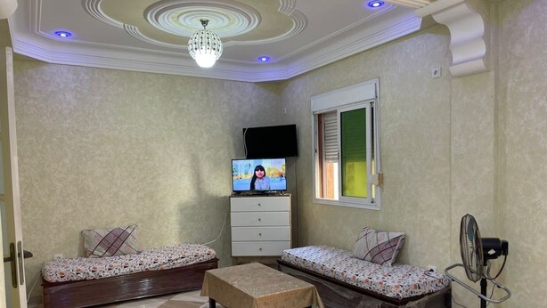 A small, well-lit room with a tiled floor, two beds with patterned bedding, a TV on a dresser, and a standing fan. - Apartment Aloue for a better stay with us welcome (Café chergui)