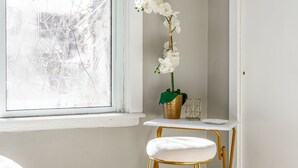 A corner room with a white chair, a small table with a white vase, and a window with a view of bare trees. - Brady St Base, 2BR Flat, Comfortably Sleeps 5 (Milwaukee)