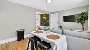 A modern living room with a grey sofa, a dining table with two chairs, a black trash bin, and a potted plant. - Brady St Base, 2BR Flat, Comfortably Sleeps 5 (Milwaukee)