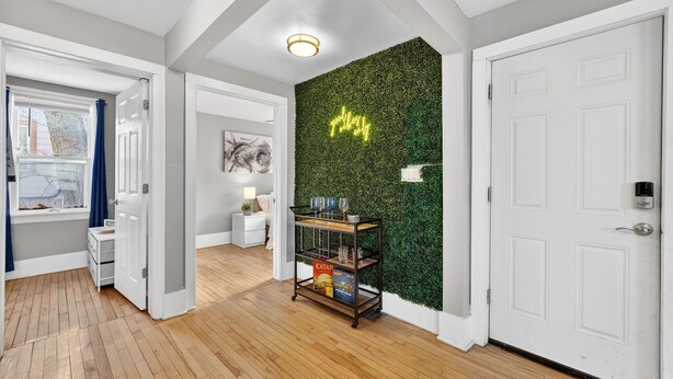 A hallway with a wooden floor, white walls, a green wall with a neon sign, and a white door. - Brady St Base, 2BR Flat, Comfortably Sleeps 5 (Milwaukee)