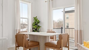Uma sala de jantar com mesa redonda, quatro cadeiras, um vaso de planta e uma varanda com vista para um edifício.
