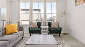 Uma sala de estar moderna com um sofá cinzento, duas poltronas verdes, uma mesa de centro branca e janelas amplas com cortinas.