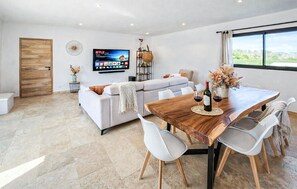 Ein modernes Wohnzimmer mit einer Couch, einem Esstisch, Stühlen, einem Fernseher und einem großen Fenster.