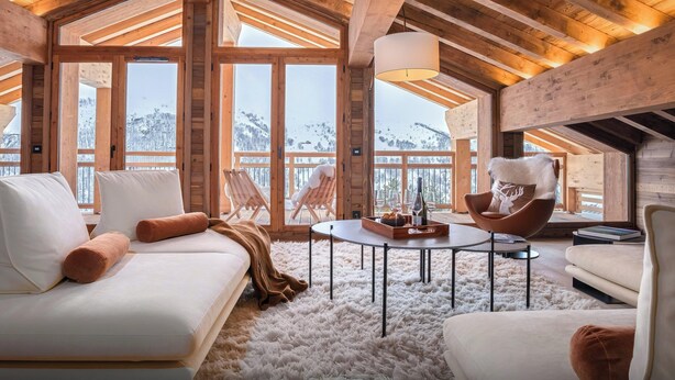 Ein gemütliches Wohnzimmer mit Holzboden, einem weißen Sofa, einem flauschigen Teppich und Blick auf verschneite Berge durch große Fenster.