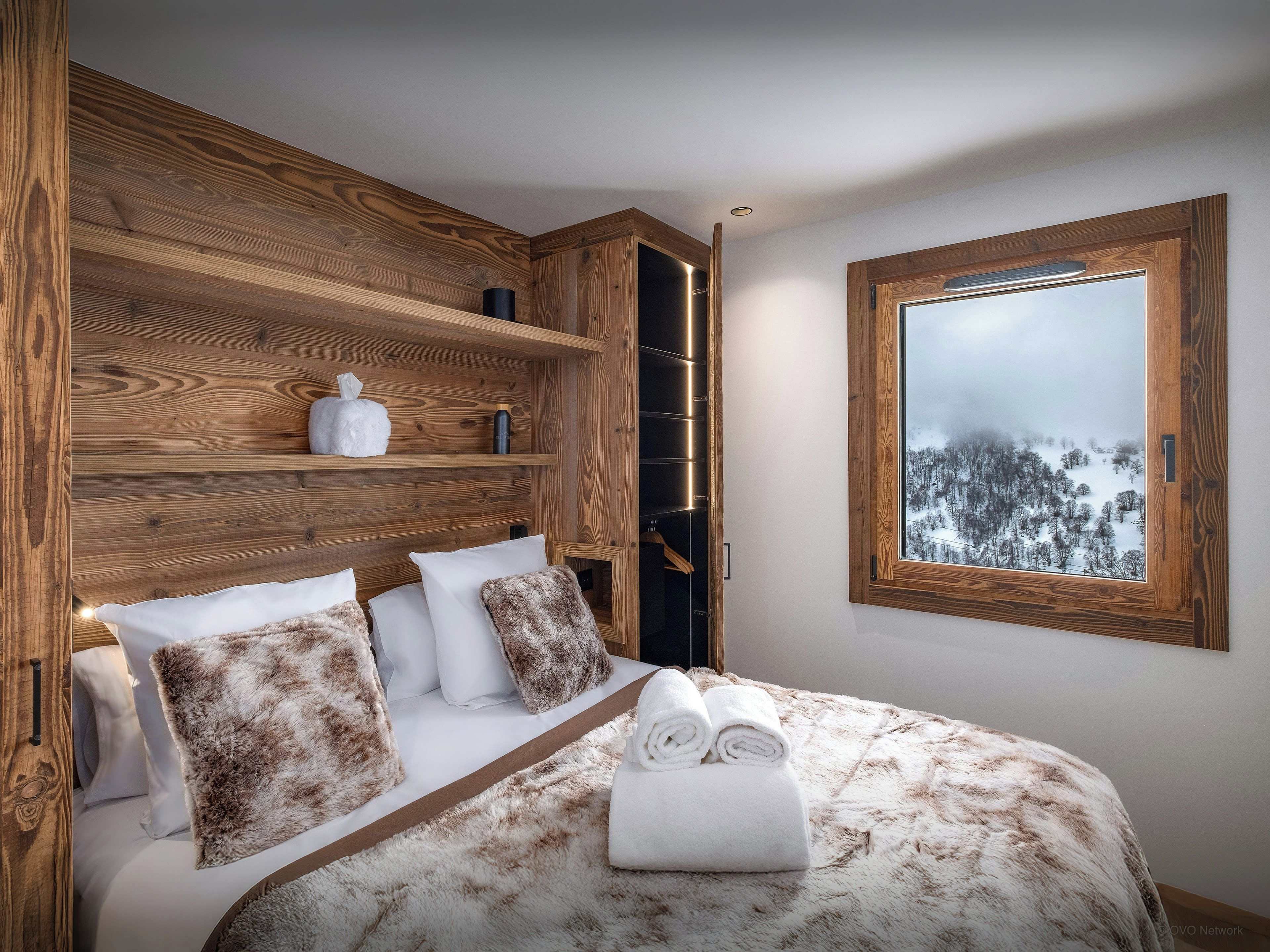 Une chambre avec une tête de lit en bois, un lit avec une literie blanche et marron, une fenêtre donnant sur un paysage enneigé et une étagère avec des articles de toilette.