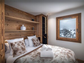 Une chambre avec une tête de lit en bois, un lit avec une literie blanche et marron, une fenêtre donnant sur un paysage enneigé et une étagère avec des articles de toilette.