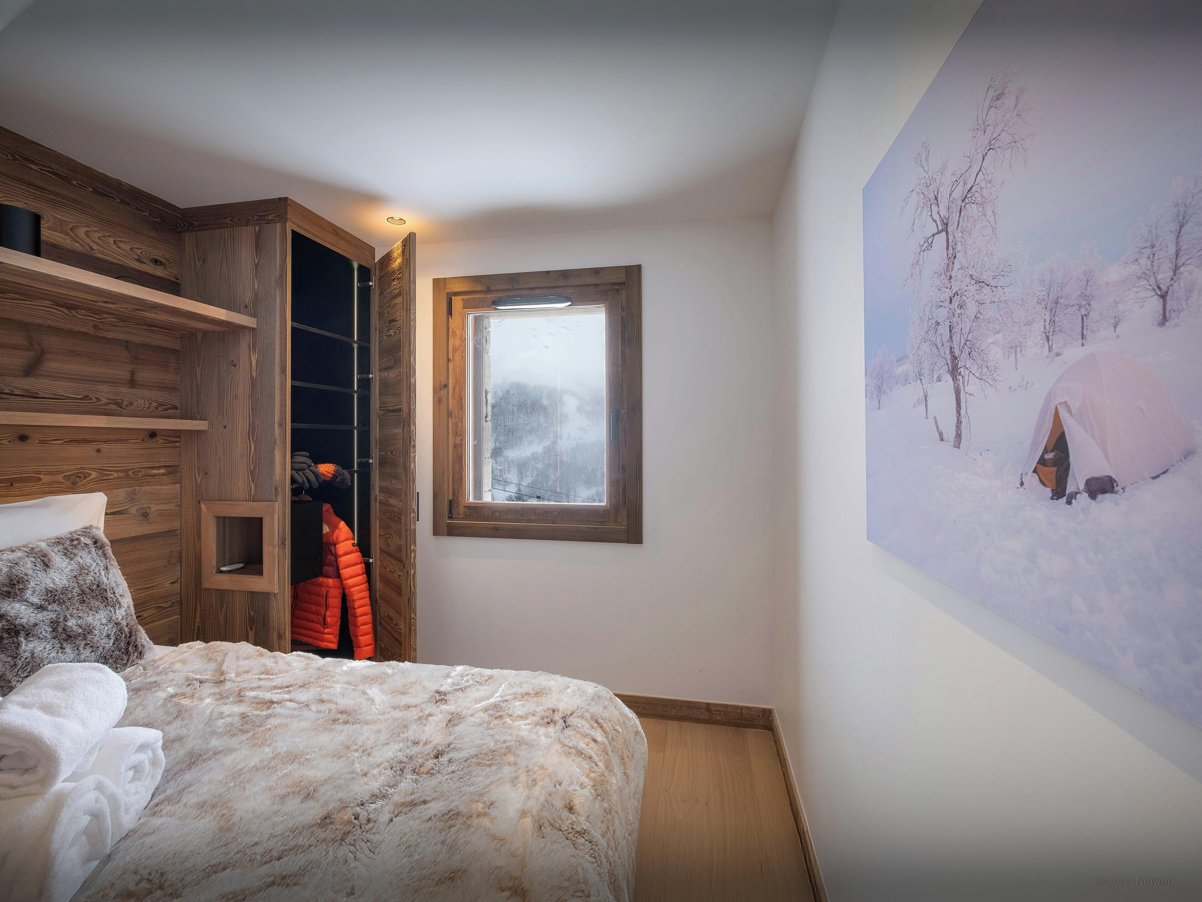 A bedroom with a wooden wardrobe, a bed with a fluffy blanket, a window with a view of a snowy landscape, and a wall with a painting of a tent in the snow.