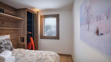 A bedroom with a wooden wardrobe, a bed with a fluffy blanket, a window with a view of a snowy landscape, and a wall with a painting of a tent in the snow.