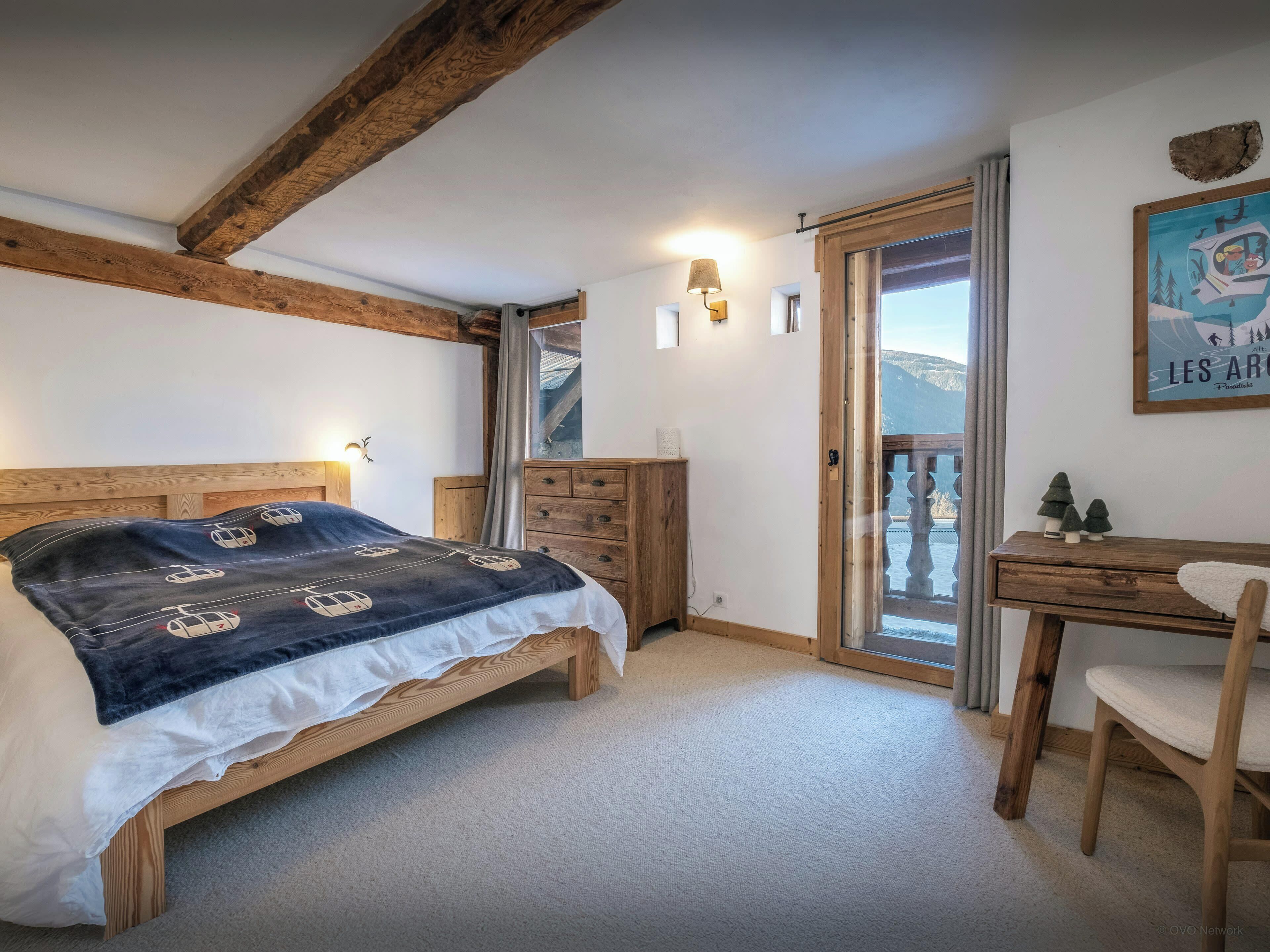 Un dormitorio con una cama de madera, un escritorio y una silla. Se ve una ventana con vistas a montañas.
