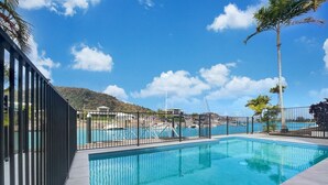 Piscina retangular com deck ladrilhado, cercada por uma grade de metal preta, e um céu azul claro acima.