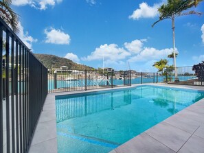 A rectangular swimming pool with a tiled deck, surrounded by a black metal fence, and a clear blue sky above. - Solmar At The Waters Edge (Nelly Bay)
